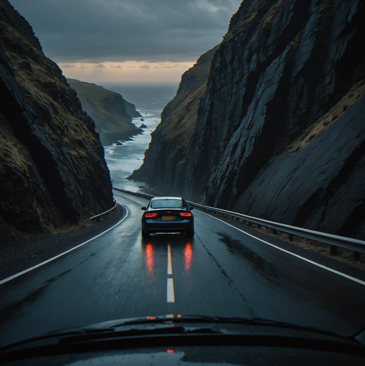 Driver's view approaching a banked cliff corner with ocean visible below, wet patch at apex creating darker asphalt reflection